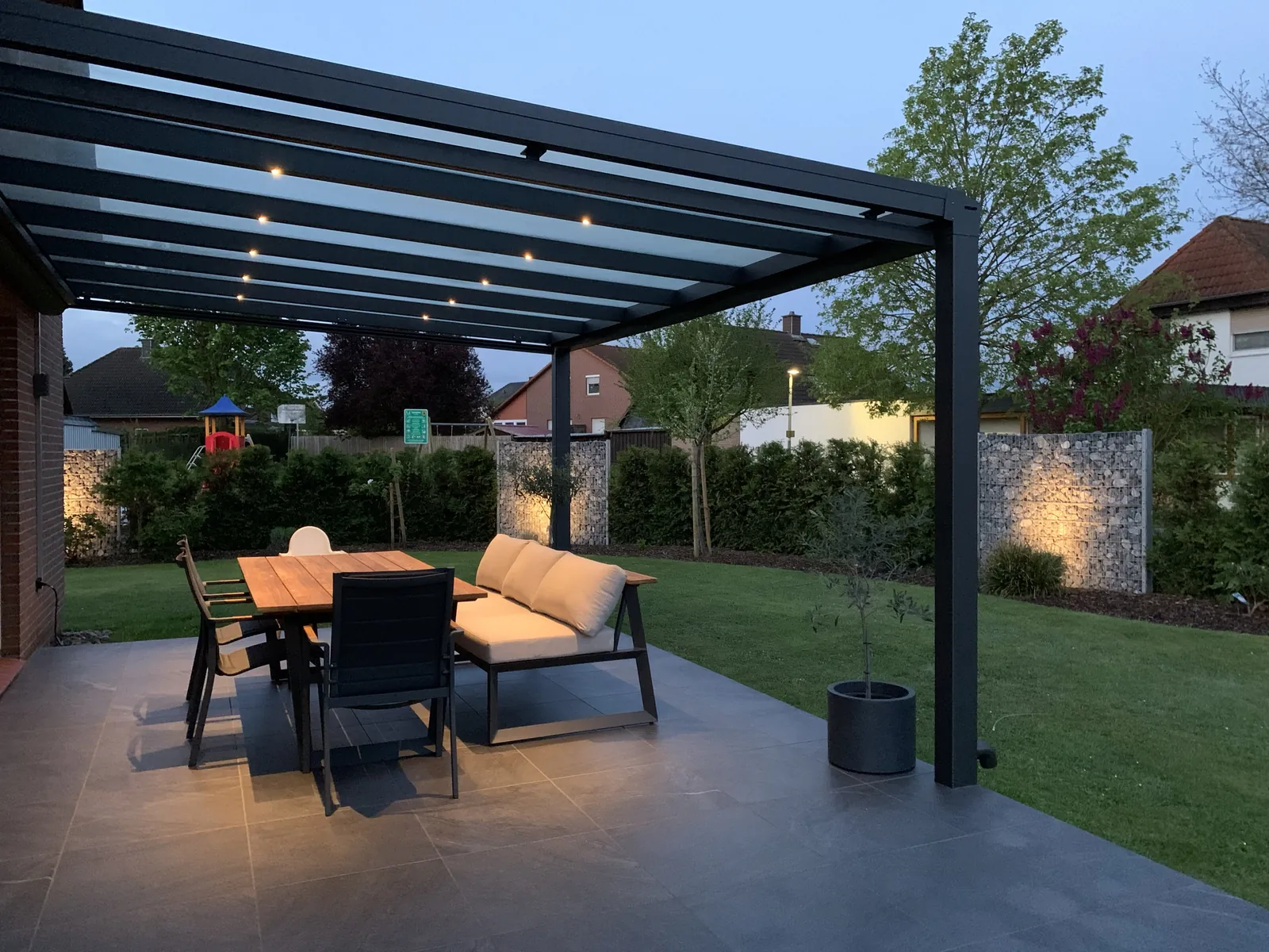 Anthrazitfarbenes Terrassendach mit Glas und LED-Spots am Abend, darunter Esstisch und Lounge